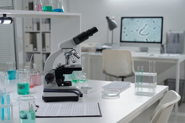 Modern laboratory setup featuring high-tech microscope and various chemical solutions on workbench. Background includes organized workspace with computer showing magnified cellular image