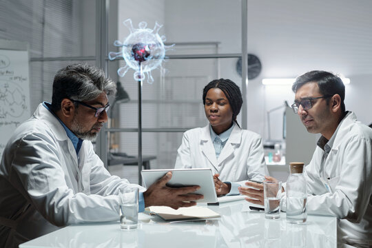 Group of scientists in lab coats collaborating around a table, sharing ideas and data on a digital tablet with holographic virus model in background