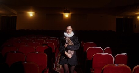 A woman with a dog in her arms stands in the aisle between the chairs in an empty theater hall. She looks at the camera with an arrogant look. © m-art