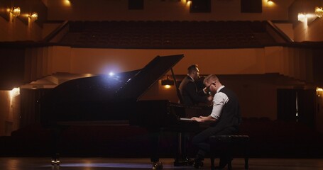 A stylish man plays the saxophone while standing on stage. The second man is playing the piano