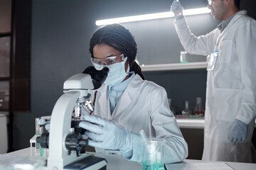Scientist conducting research with microscope while colleague working in background, wearing lab...