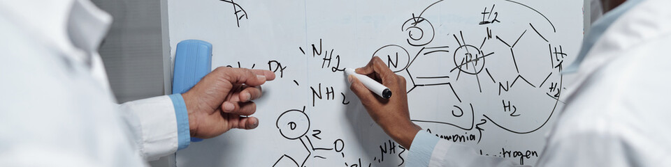 Two individuals drawing complex chemical structures on whiteboard with detailed notations. Wearing...