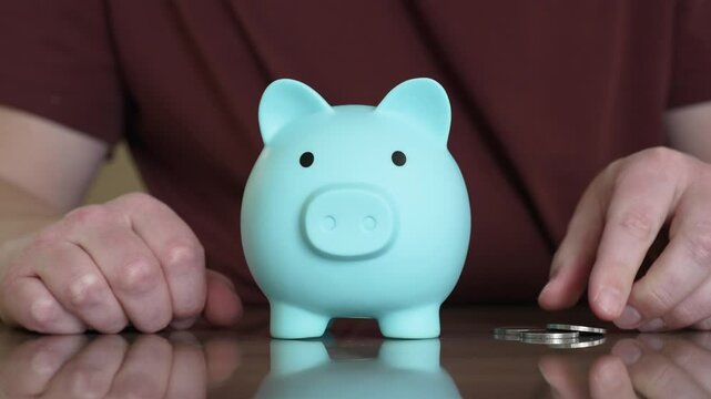 Placing coins into light blue piggy bank positioned on shiny surface, visualizing systematic money saving and financial growth strategy