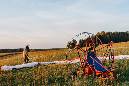 Paraglider man lays out soft self-inflating wing before the flight, preparing to fly on red paraplane in sky above fields in countryside in summer at sunset