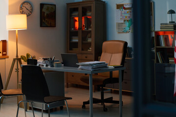 Lamp standing by wall of office with workplace of school psychologist consisting of armchair and desk with documents and laptop