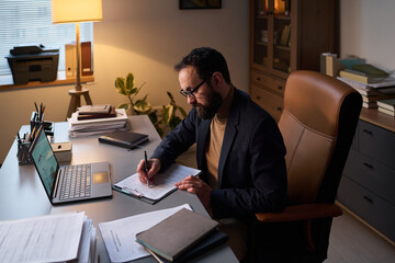 Bearded man in formalwear and eyeglasses bending over his workplace while sitting in office and...