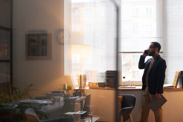 Side view of mature school psychologist with clipboard in hand speaking on mobile phone while standing by office window