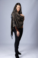 A  dark-haired woman wearing black pants, a brown top and a woven paisley shawl draped over her...