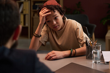 Sad schoolboy touching his head and looking at mature school psychologist during discussion of his problems