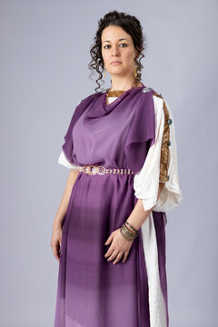 A high-status Roman woman wearing a white tunic with a purple silk stola over the top. She had dark, curly hair and stands against a studio backdrop. 