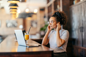 Young interracial female freelancer is putting earbuds and preparing for conference call on laptop in cafe.