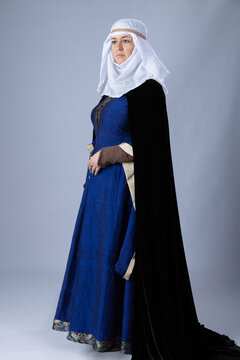 A Medieval woman wearing a blue silk gown called a bliaut and a white linen wimple against a plain studio backdrop