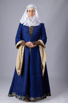 A Medieval woman wearing a blue silk gown called a bliaut and a white linen wimple against a plain studio backdrop
