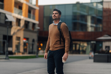 Obraz premium Young businessman walking in the city holding a coffee and wearing a backpack