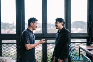 Two young men in casual wear chatting, near window with cityscape background