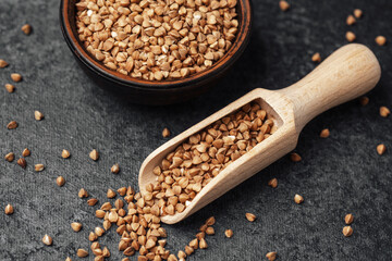 Buckwheat grains in a wooden scoop with a bowl on a dark surface