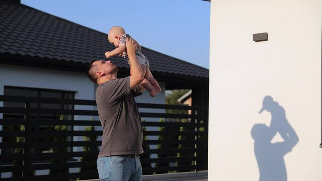 A joyful young father lifts his child into the air and kisses him while standing in a landscaped yard. Warm sunlight casts playful shadows against a modern house.