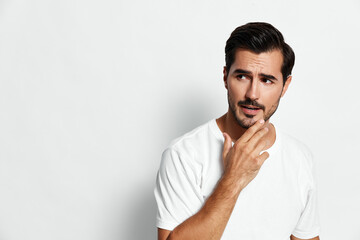 Thoughtful young man in white t-shirt touching his beard, pondering, looking sideways with a...