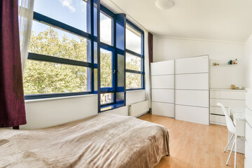 A spacious and well-lit bedroom featuring large windows, white furniture, and a cozy bedding arrangement. The design emphasizes simplicity and modernity.
