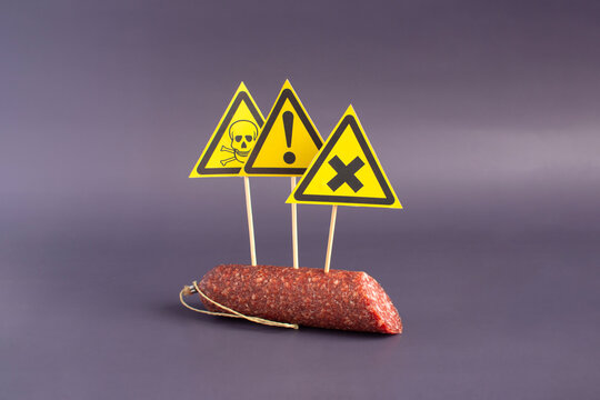 The concept of dangerous sausage. A fragment of smoked sausage and warning signs in the form of yellow triangles in close-up on a dark background.