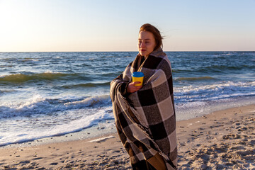 Woman wrapped in blanket holding coffee on windy beach