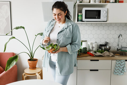 Happy plus size woman in casual clothes holding transparent bowl of delicious tasty fresh organic salad, mixing it with spoon standing at kitchen, losing weight and trying to stay healthy