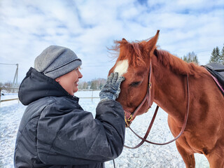 Woman interacts affectionately with a chestnut horse during a winter day at the stable in a snowy...