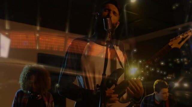 bandmates performing music under warm lights, singing into mic and strumming guitars with city glow
