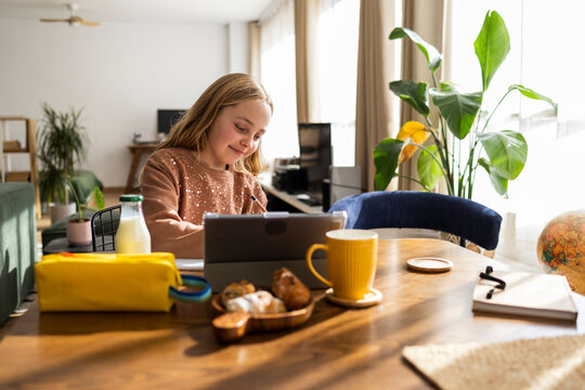 Girl doing homework on a tablet in a cozy living room with breakfast on the table