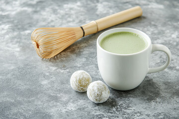 creamy matcha latte in white mug sits beside two sweet mochi treats, with bamboo whisk in background, creating serene and inviting atmosphere for tea lovers