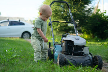 Fototapeta premium Young toddler carefully studying lawn mower in grassy yard, learning about outdoor equipment. Concept of safe play areas and proper lawn care.
