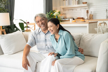 Obraz premium Asian senior couple laughing and smiling together on couch. Retired grandparent relaxing at home in the morning. Healthy elderly people and life insurance concept