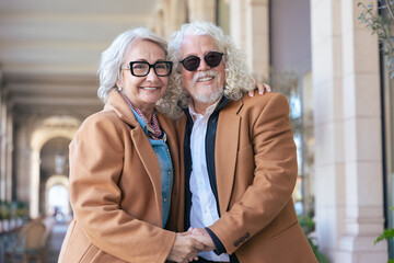 Happy caucasian elderly couple standing close, smiling and looking at the camera. Dressed in brown coats, they pose affectionately in a sunlit outdoor corridor .