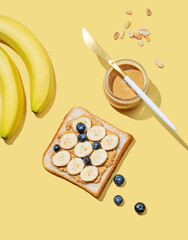 Peanut butter toast with banana, blueberry, nuts and knife on a yellow background.