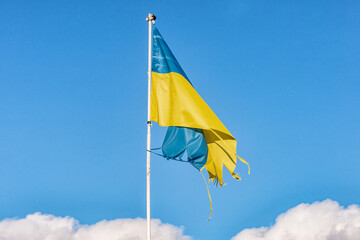 A tattered Ukrainian flag fluttering in the wind against a blue sky with clouds. The photo symbolizes resilience, struggle, and unwavering faith in victory.