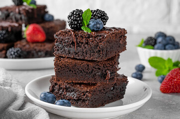 A stack of chocolate brownies with chocolate glaze, fresh berries and mint leaves on top on a white plate on a gray concrete background. Delicious dessert.