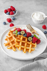 Delicious Belgian Waffle with whipped cream, fresh berries and mint on a white plate on a gray concrete background. Summer breakfast.