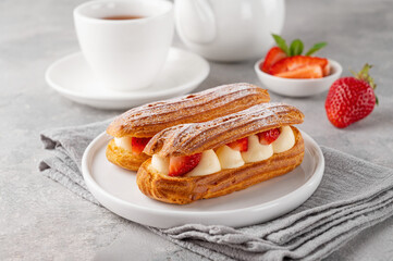 Eclairs with cream and fresh strawberries sprinkled with powdered sugar on top on a gray concrete background.