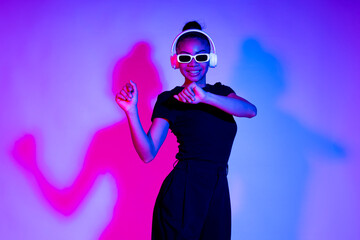 Woman dancing in a studio with neon lights wearing wireless headphones and sunglasses