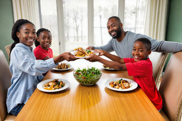 African american family enjoying dinner together at wooden table. Father and three smiling daughters passing plates with chicken, vegetables and salad during mealtime. Healthy meal with fresh food
