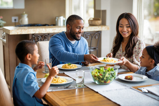 Multiracial family sharing pasta dinner at wooden table. Father in blue sweater passes plate to mother in floral blouse while children watch. Healthy meal with salad, homemade food, and happy
