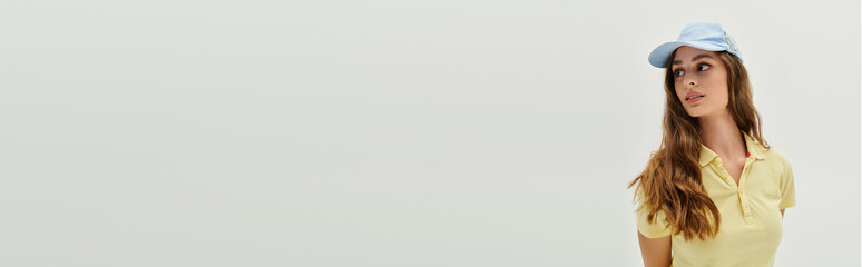 Beautiful young woman wearing a yellow shirt and cap poses gracefully against a plain background