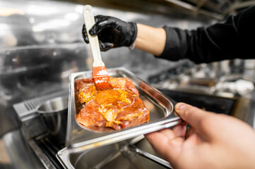 A chef in a black glove brushes marinade onto a piece of raw meat in a metallic tray. The kitchen environment reflects a busy, professional setting focused on culinary preparation.
