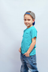 Smiling child wearing a backward cap and turquoise t-shirt with hands in jean pockets, standing in profile. The child has light brown hair and a relaxed, confident expression