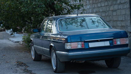 Classic blue sedan parked on a quiet street with lush greenery nearby during evening hours