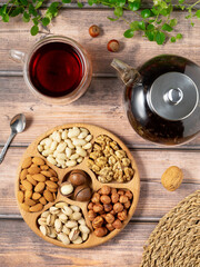 A high-angle flat lay shot showcasing a healthy assortment of nuts and a hot beverage on a rustic, light brown wooden surface. The central element is a round wooden serving tray with six compartments,