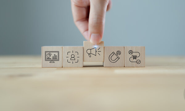 Marketing strategy planning with wooden blocks. A hand arranging wooden blocks with  icons representing data analytics, customer targeting, advertising, lead generation and sales process.