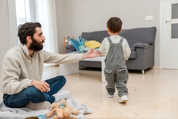 Dad sitting on the floor watching his baby son walking in living room