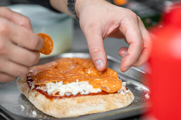 A chef's hands place slices of pepperoni on a pizza topped with cheese and tomato sauce on a metallic tray. The vibrant kitchen setting reflects an atmosphere of culinary creativity.
