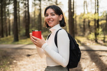 Woman hiker in casual gear sipping from a thermo mug in a peaceful forest, merging travel lifestyle with wellness vibes